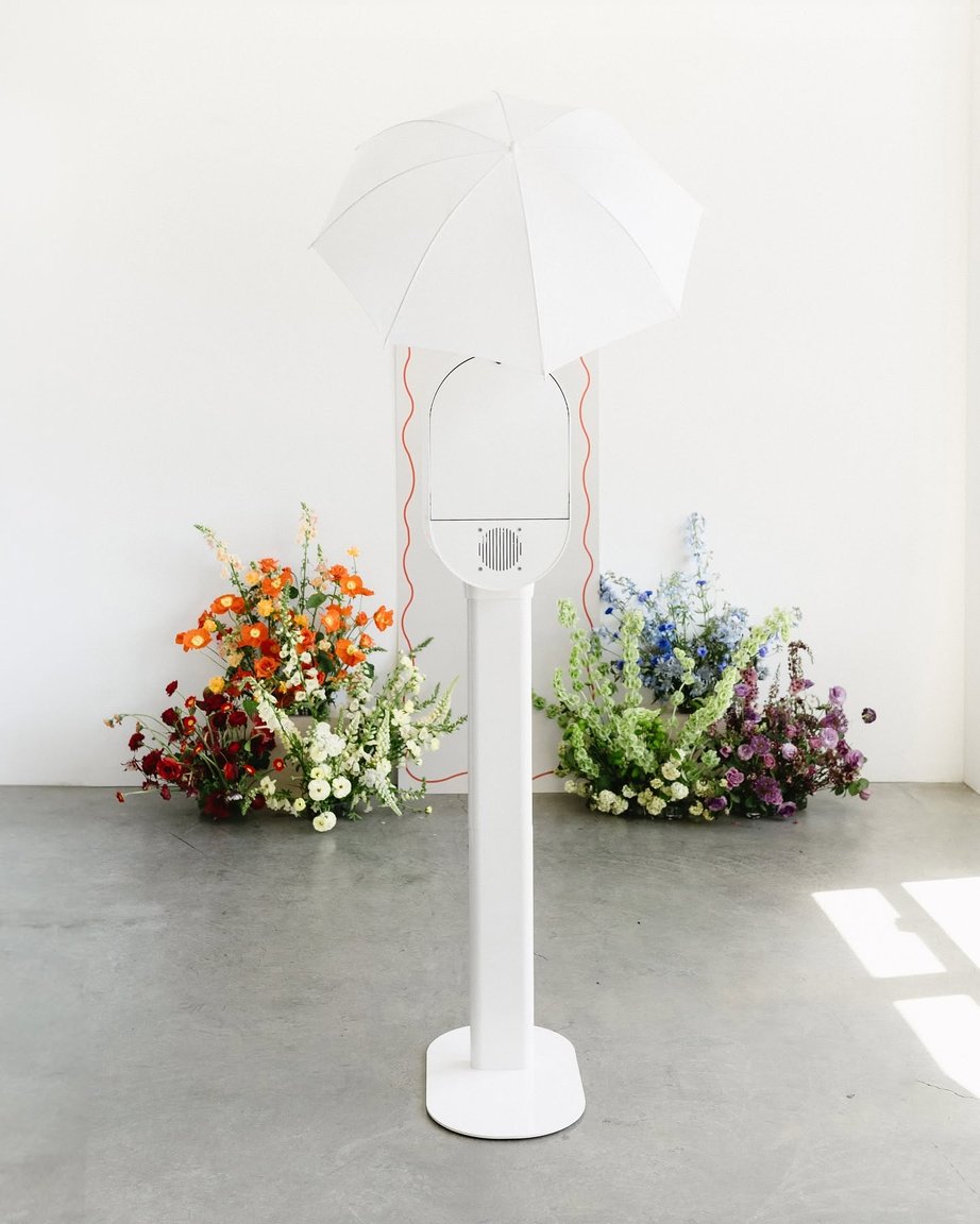 The white photo booth and its umbrella light between two flower arrangements.