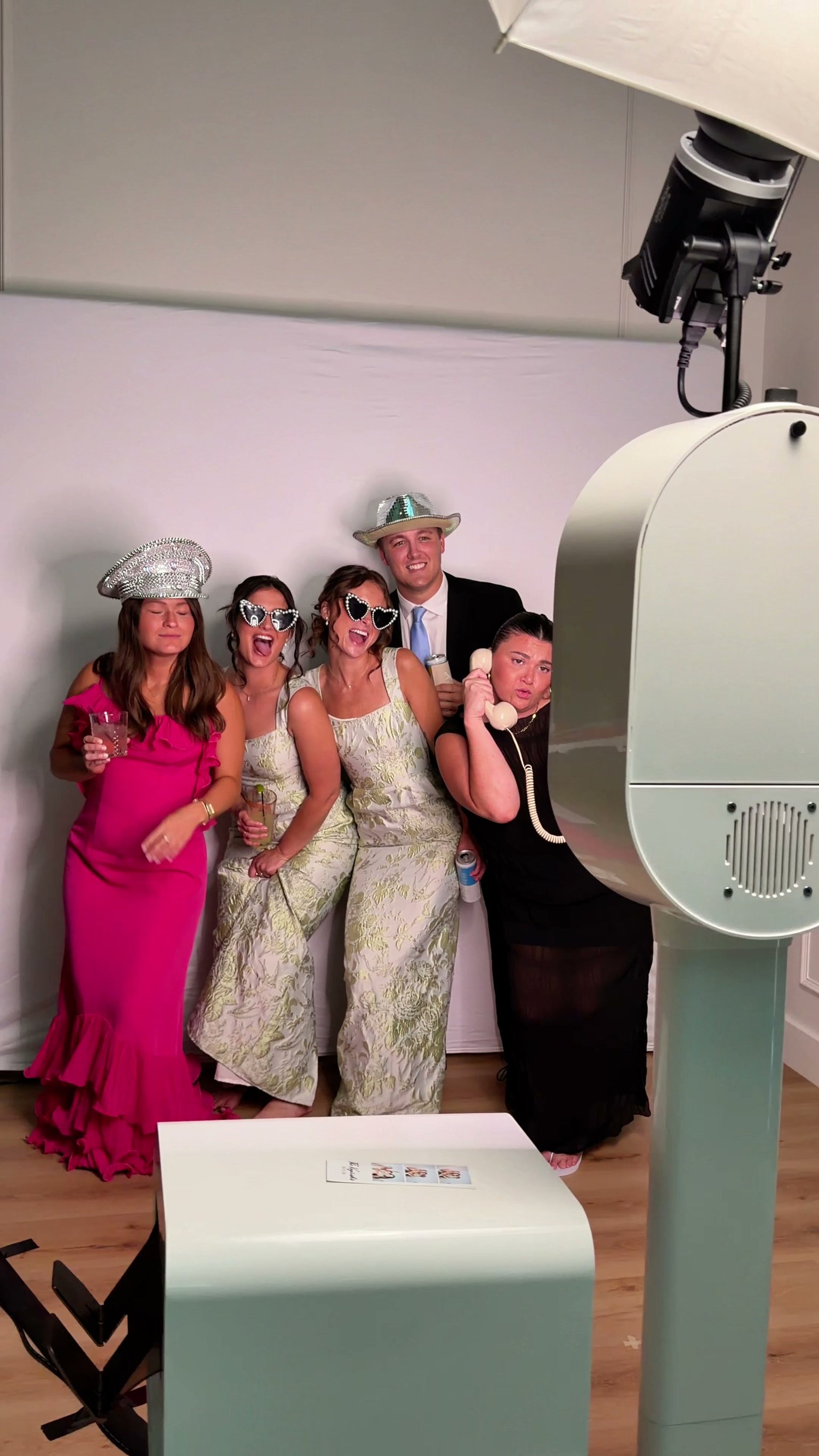 Five guests in props posing at the photo booth, one on the retro phone, with the studio light above and the printer in front.