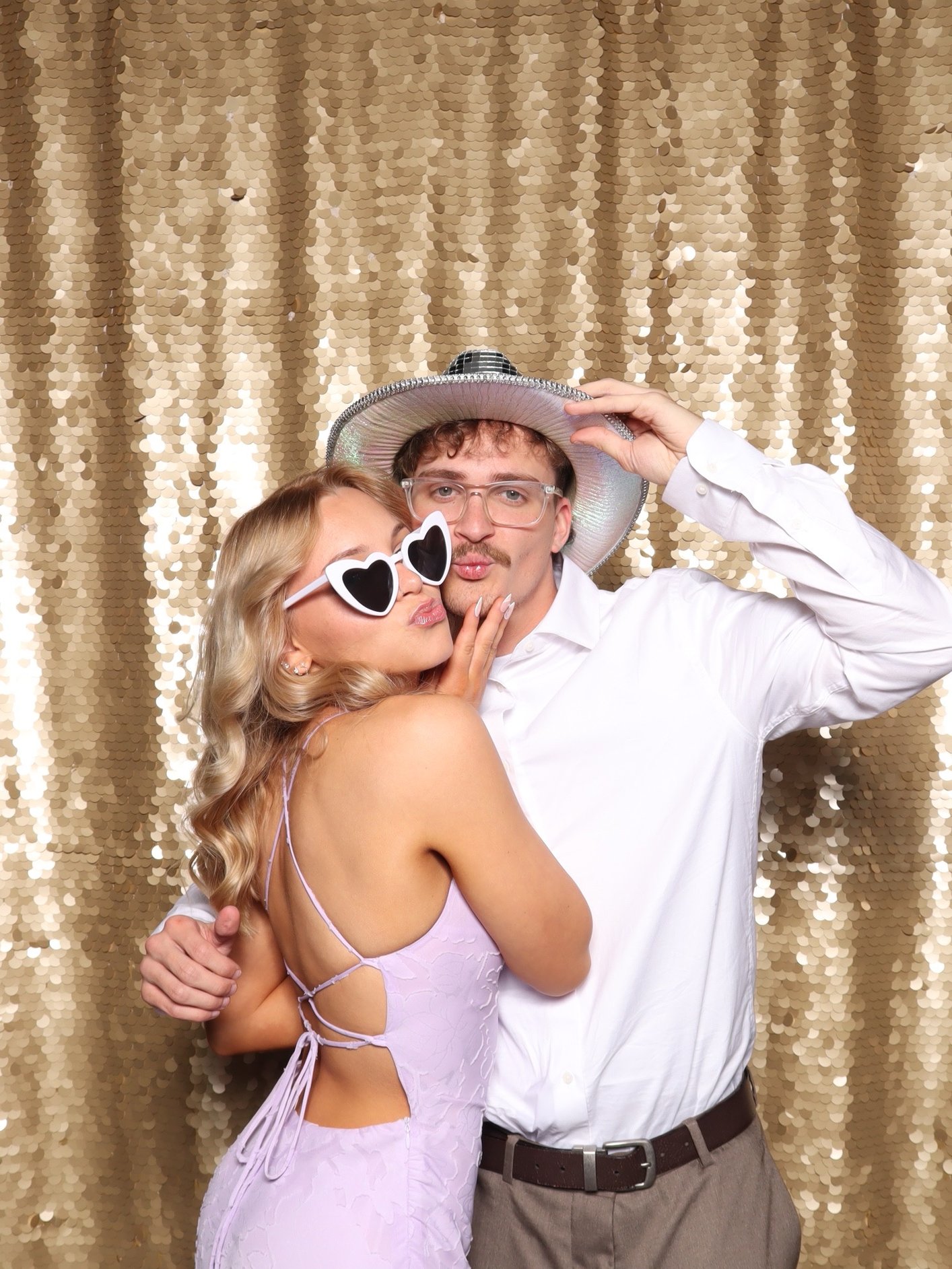 A couple in heart sunglasses and a disco cowboy hat at a gold sequin backdrop.