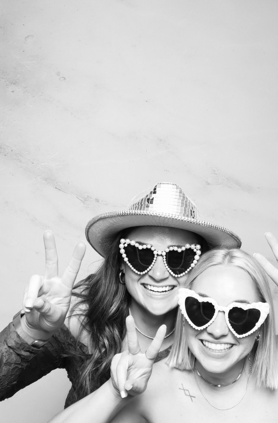 Two friends in heart sunglasses, one wearing a disco cowboy hat, at the photo booth.
