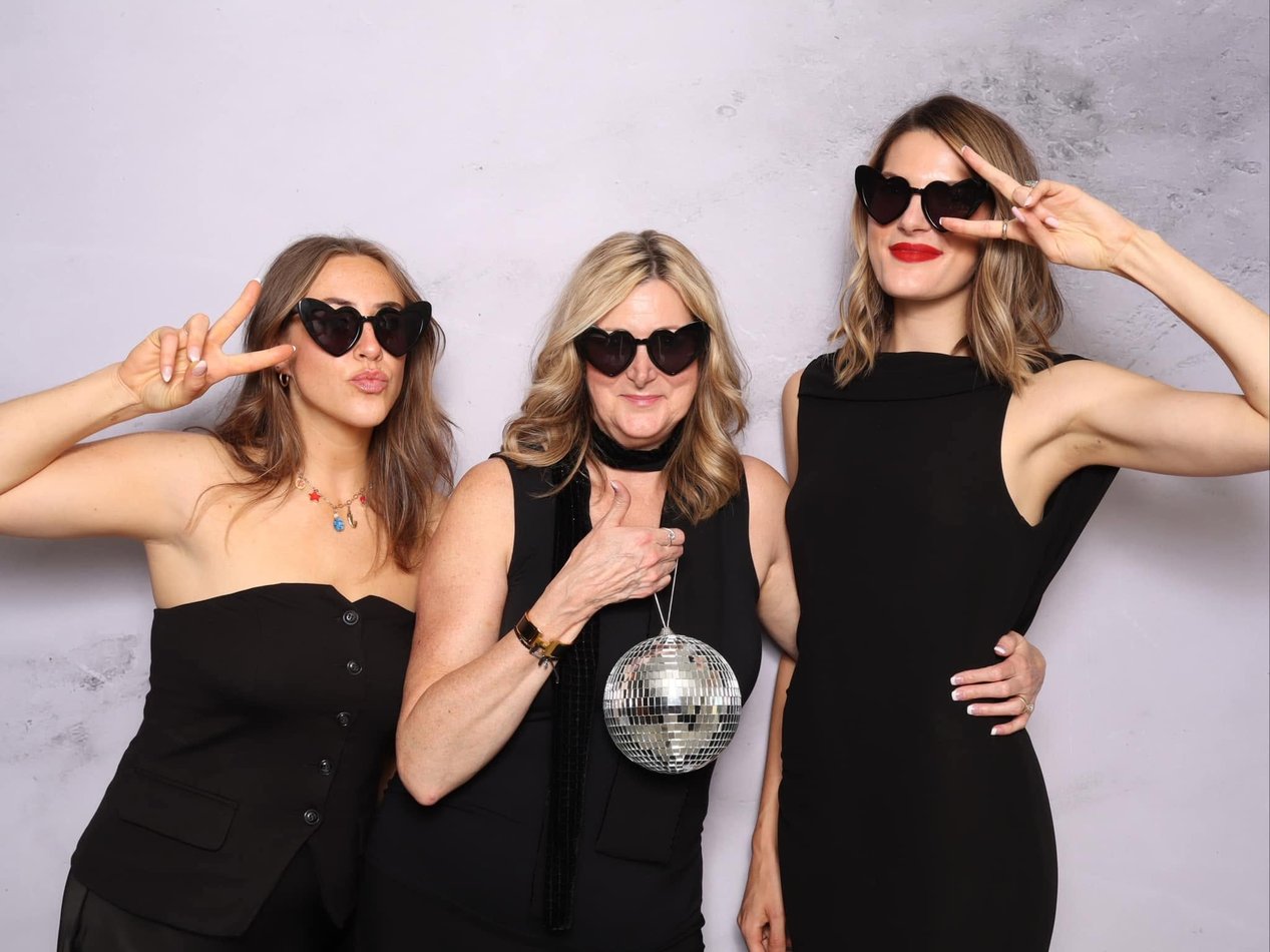 Three women in heart sunglasses, one holding a disco ball, at the photo booth.